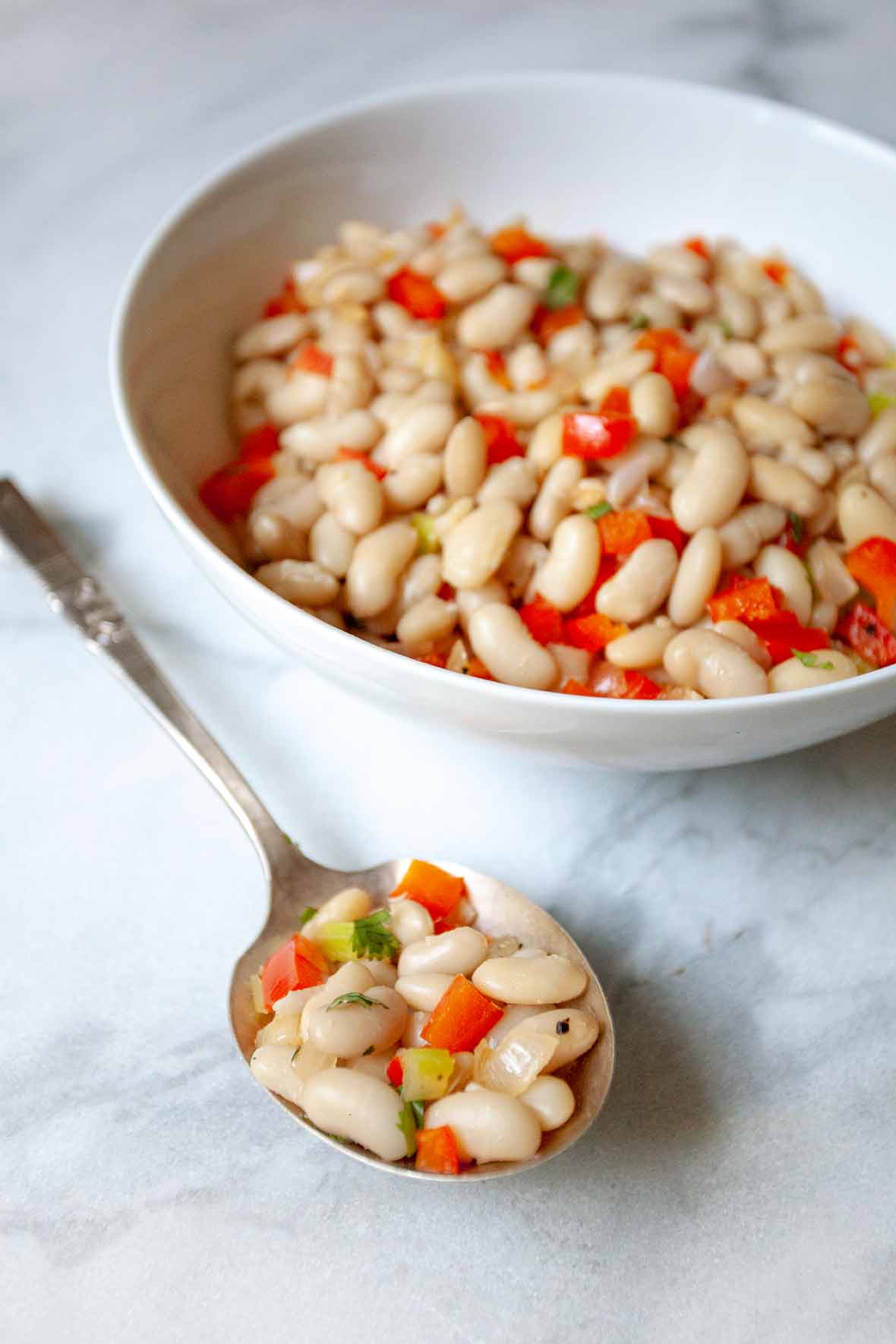 A white bowl and silver spoon filled with tuscan beans, red pepper, and celery.