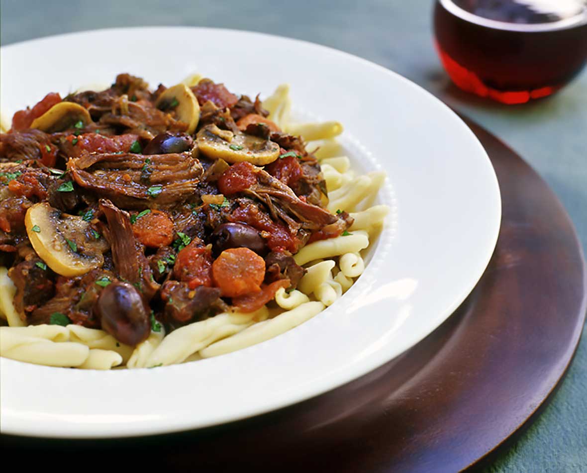 Bowl of french beef stew with sliced carrots over pasta sitting on a wooden tray