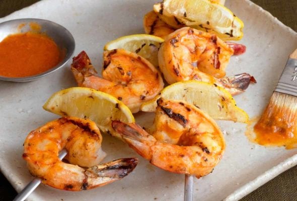 A white square platter with two skewers of grilled Portuguese shrimp with piri piri sauce and lemon wedges, a small dish of piri piri sauce, and a basting brush.