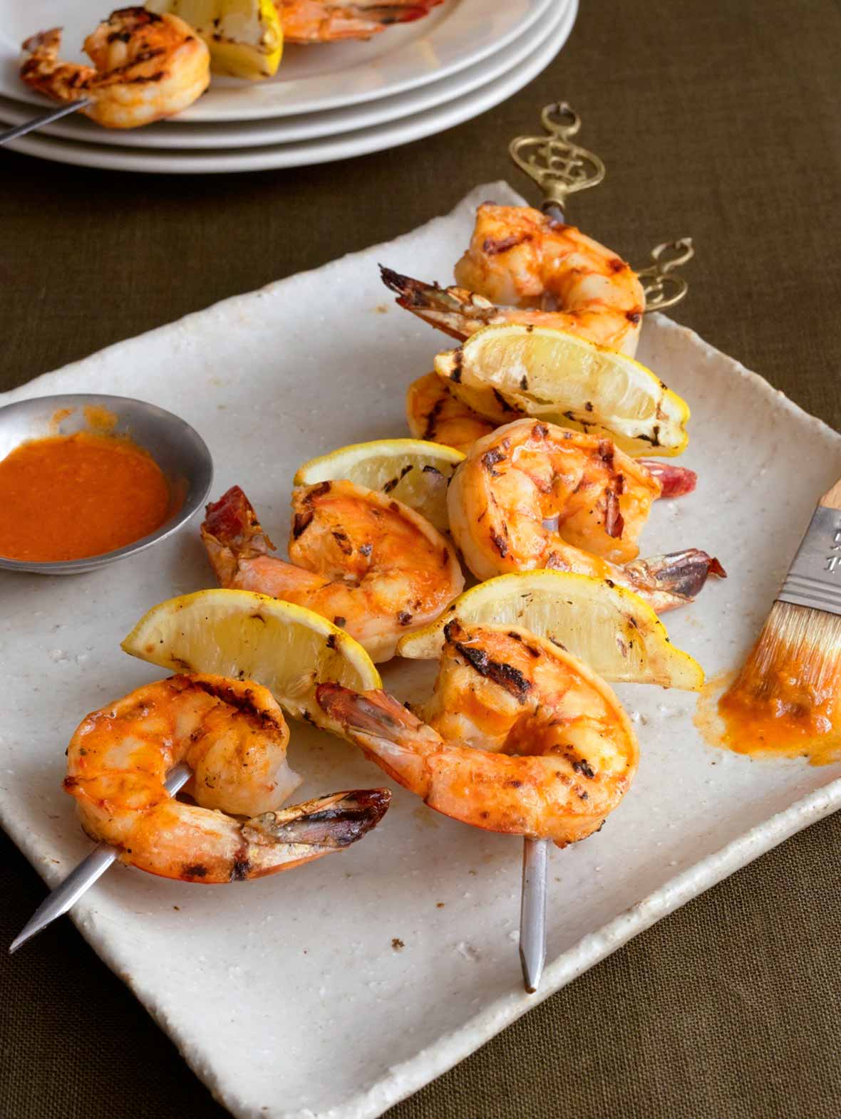 A white square platter with two skewers of grilled Portuguese shrimp with piri piri sauce and lemon wedges, a small dish of piri piri sauce, and a basting brush.