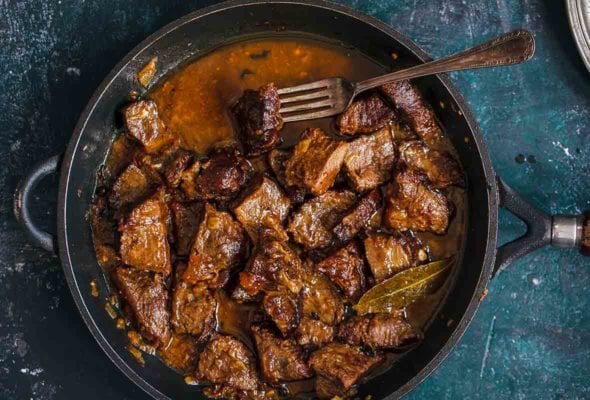 A cast iron skillet with cacoila--or chucks of Portuguese stewed beef, bay leaf, and a fork