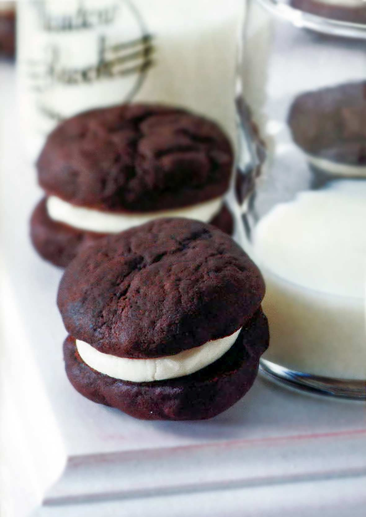 Two whoopee pies--chocolate cookie sandwiches with cream filling on a white table with a glass of milk