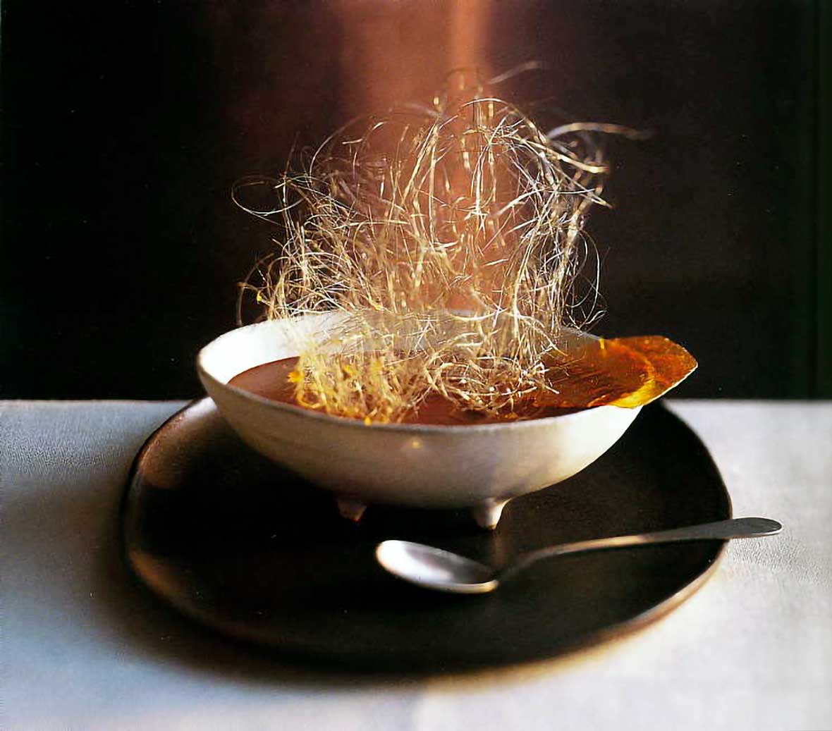 A white bowl of chocolate mousse with spun sugar on top, nearby a spoon