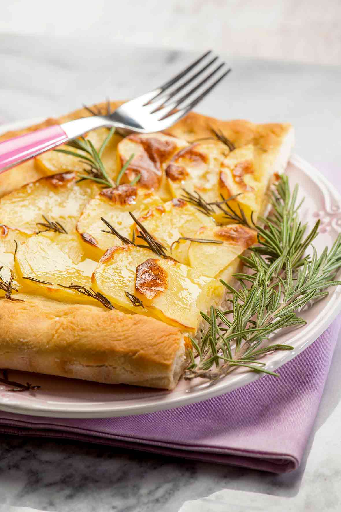 A potato rosemary focaccia--bread topped with sliced potatoes and rosemary sprigs on a white plate