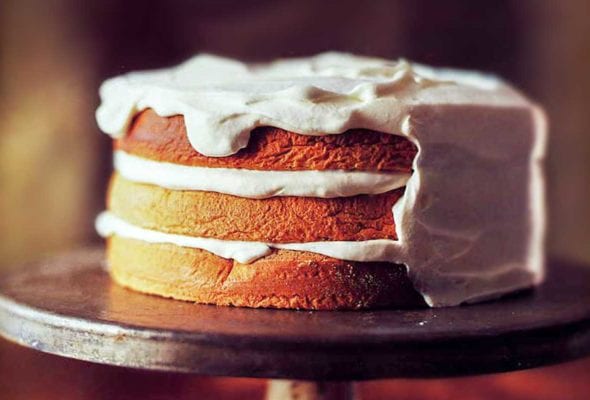 A partially frosted three-layer walnut torte on a vintage cake stand with a bowl of whipped cream and a knife beside it.