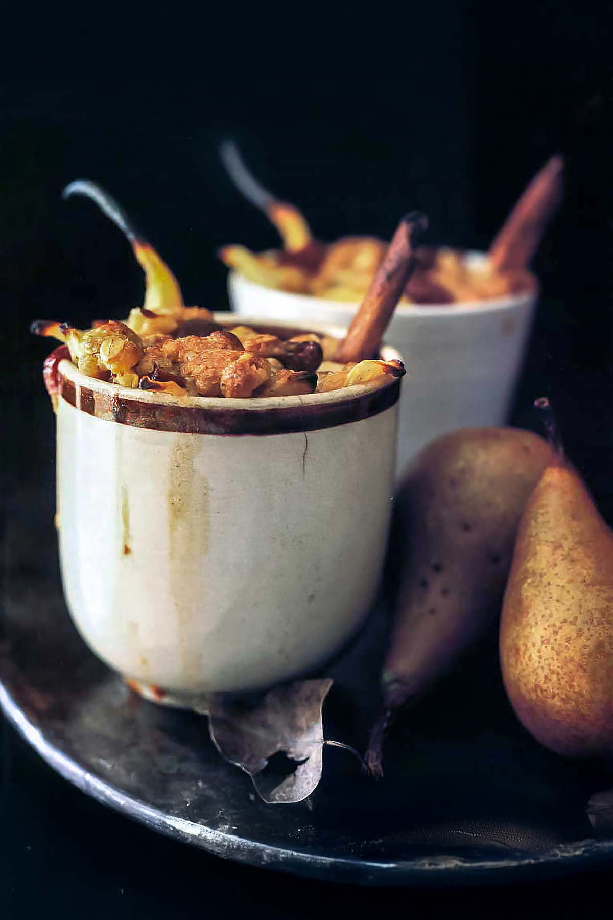 Two cup filled with pear-cranberry crumble, two pears, on a metal plate