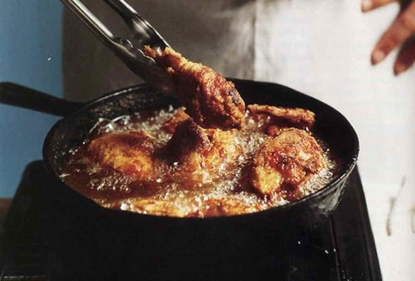 A man holding tongs in front of a skillet making southern pan-fried chicken, which is bubbling in the pan