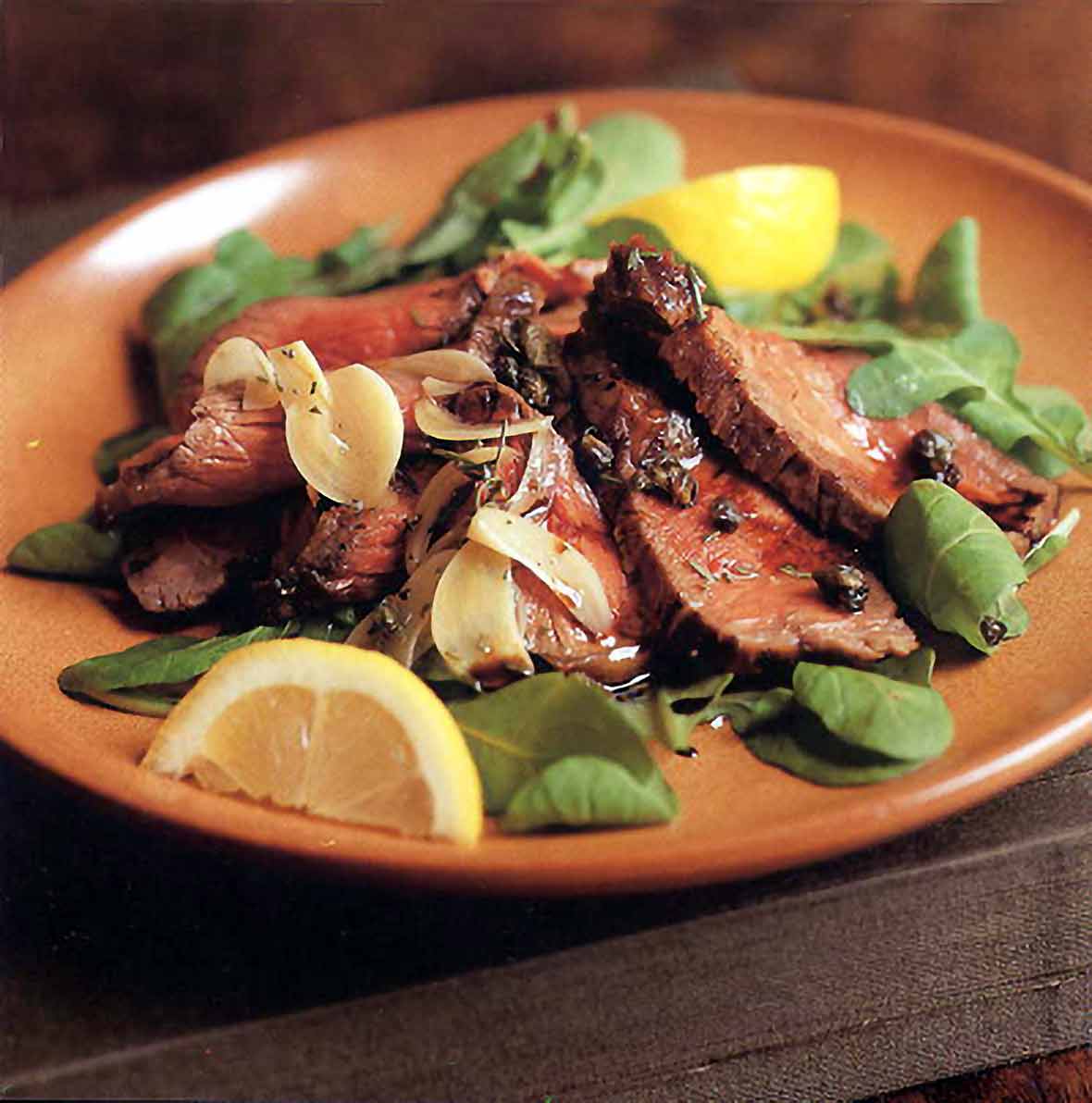 Plate of grilled beef, rosemary, capers, salad greens and lemon wedges