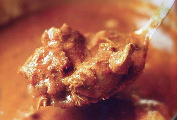 A spoon of a red chicken curry held above copper pot filled with the Indian stew