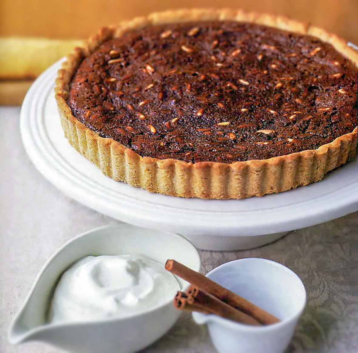 A chocolate tart with a fluted crust on a white cake stand; whipped cream and cinnamon sticks on the side