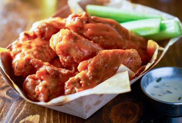A paper basket filled with classic buffalo wings and celery sticks with a metal cup of dipping sauce beside it.