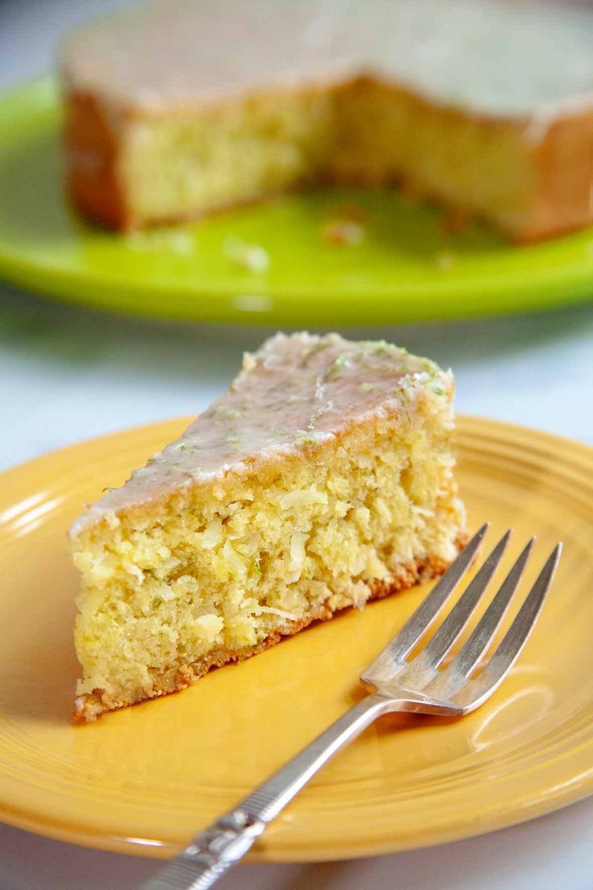 A slice of coconut lime macadamia cake on a yellow plate with a fork resting beside it