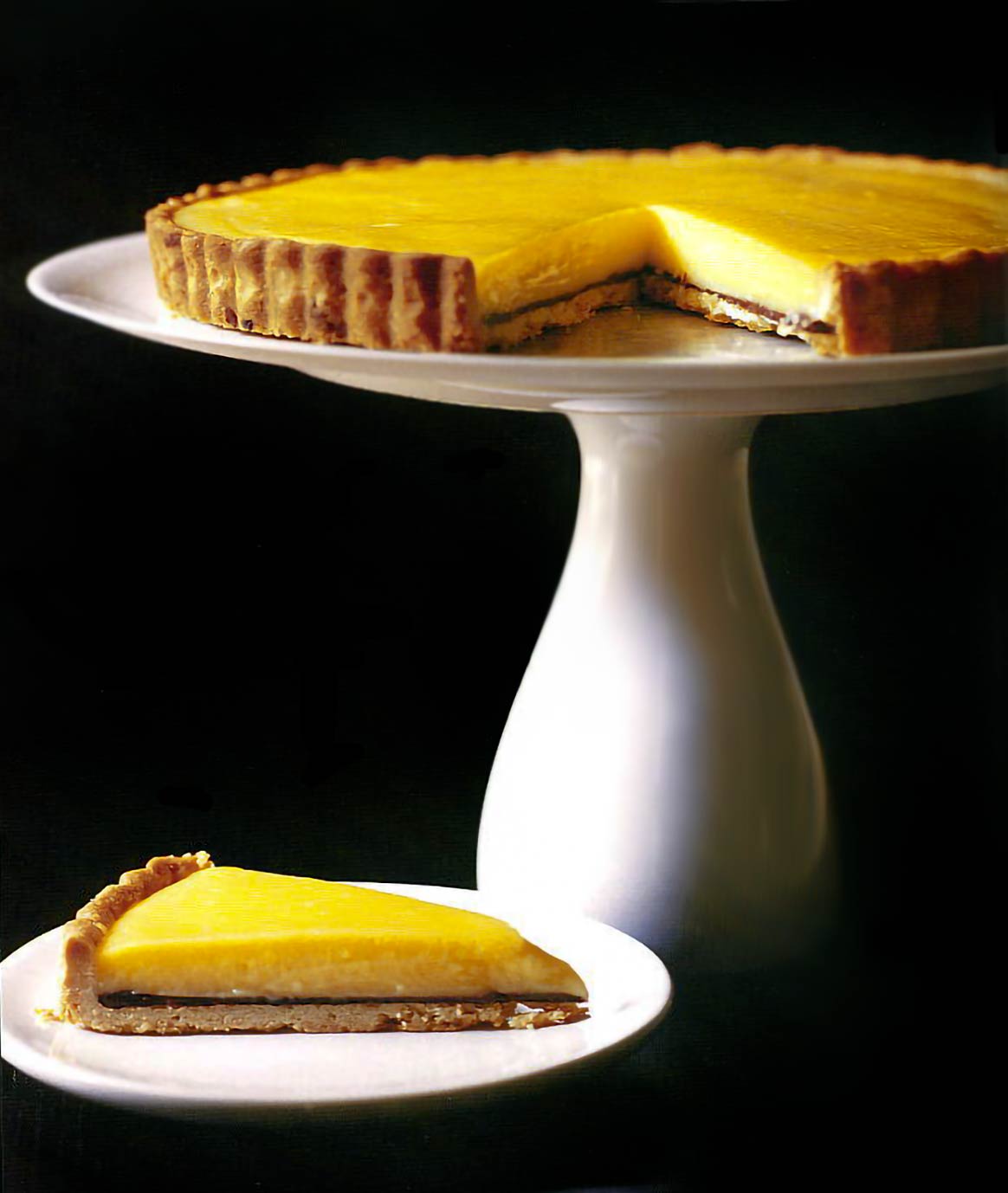 Meyer lemon tart on a white cake stand and a slice of tart on a small plate, black background