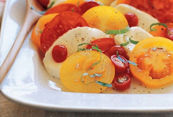 A white serving platter filled with Caprese salad made with sliced tomatoes, sliced mozzarella, and fresh basil with a serving spoon resting inside.