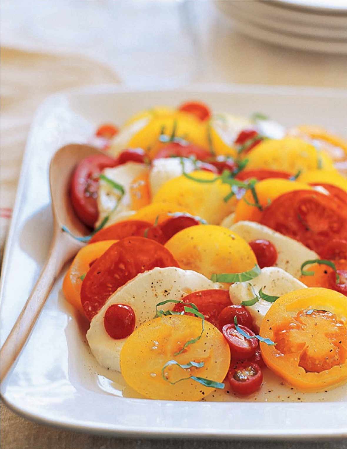 A white serving platter filled with Caprese salad made with sliced tomatoes, sliced mozzarella, and fresh basil with a serving spoon resting inside.