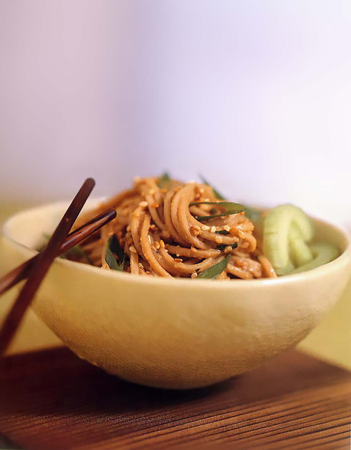 A bowl of sesame-peanut noodles coated in peanut sauce, topped with sesame seeds, chopsticks on the side