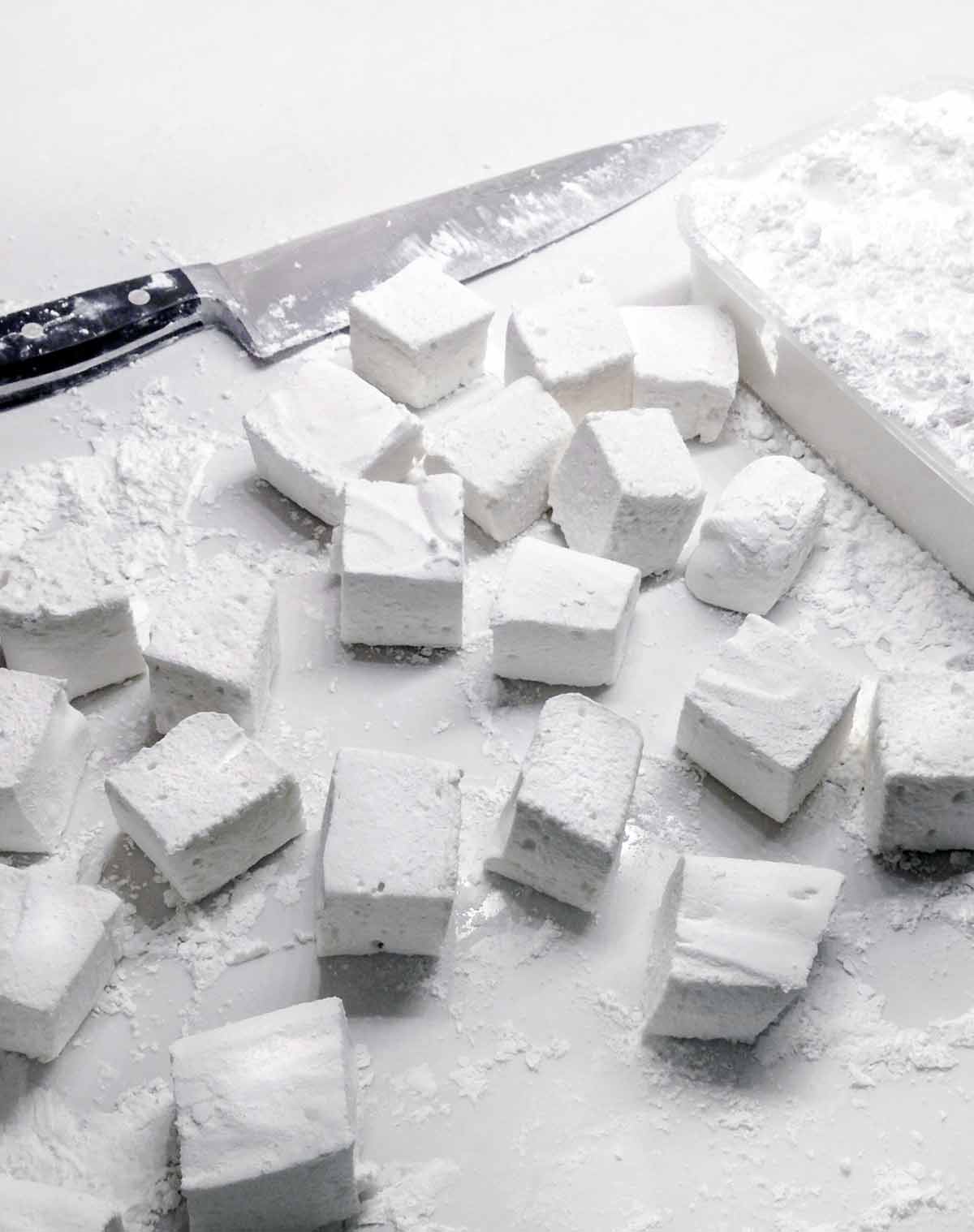 Cubes of homemade marshmallows on a white surface with a chef's knife lying beside.
