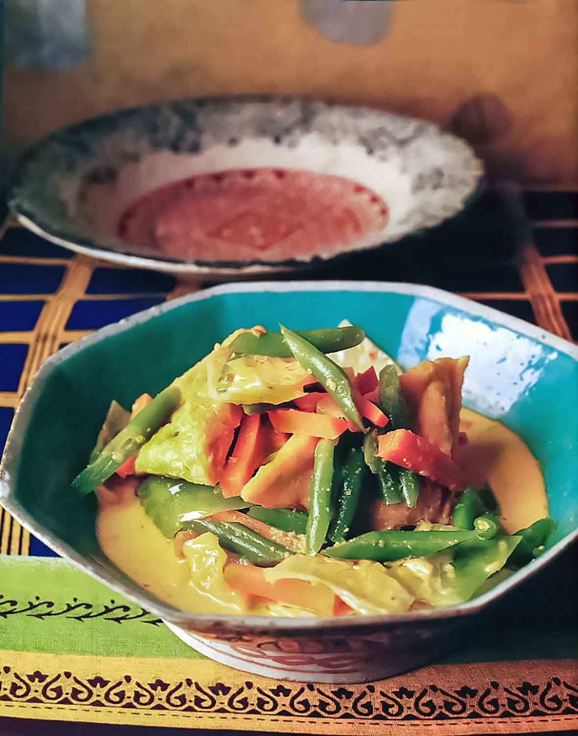 An octagonal bowl filled with tofu and vegetables in coconut milk on a colorful cloth with a large bowl in the background.