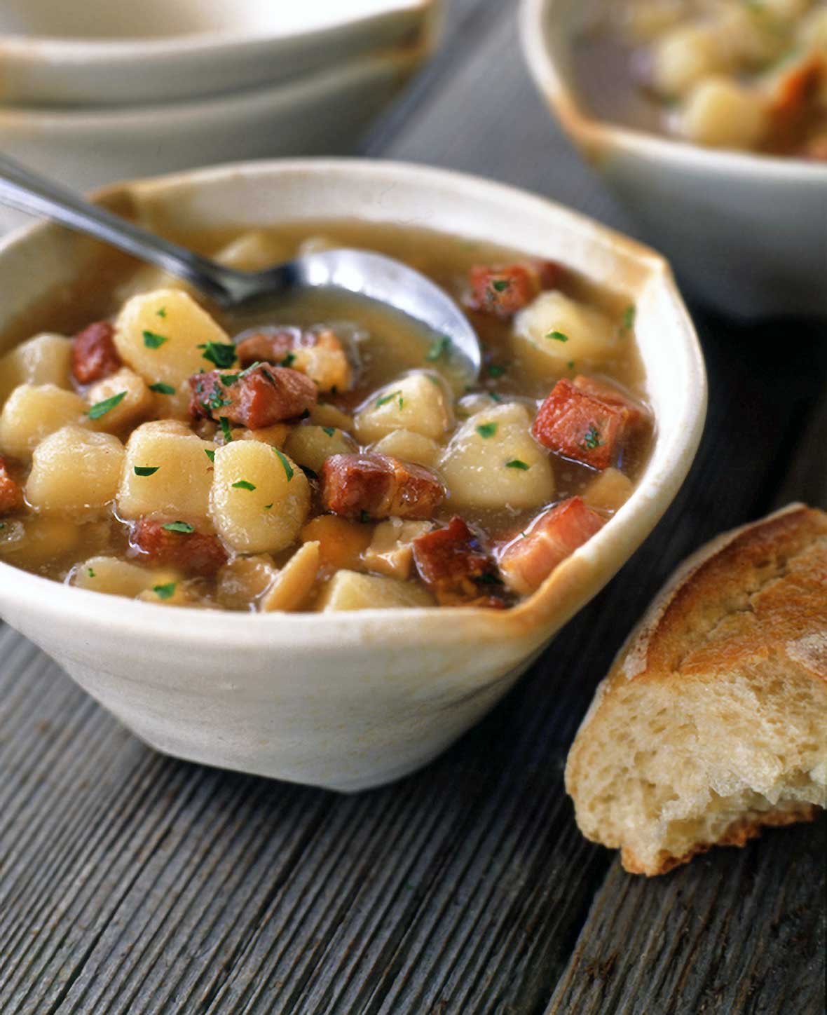 Bowl of Rhode Island clam chowder with quahogs, diced potatoes and bacon on wood, bread and bowls nearby