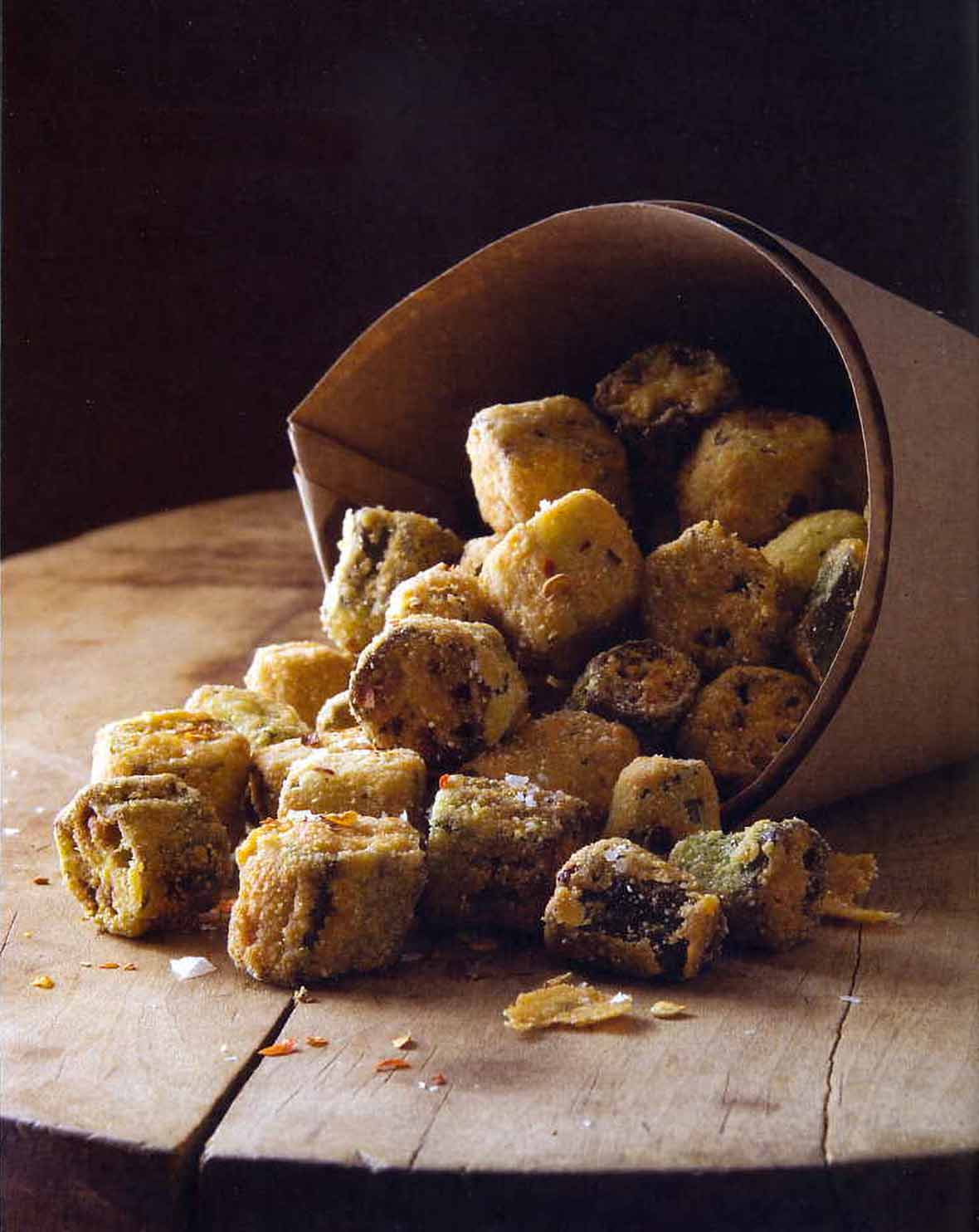Paper cone with crispy fried okra bites spilling out on top a wooden board