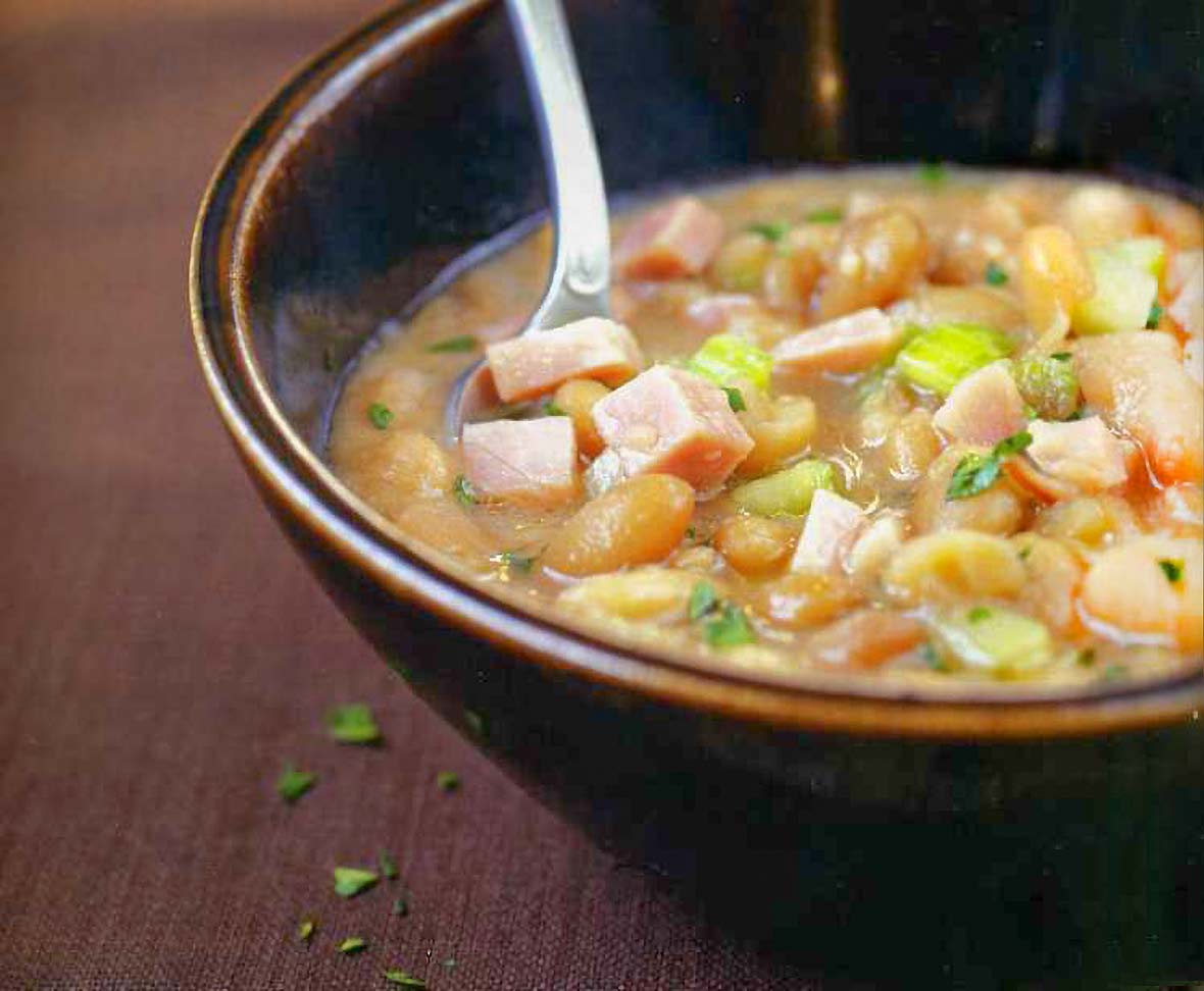 Bowl with a soup of navy beans, ham hocks, onions, celery, and parsley on brown fabric