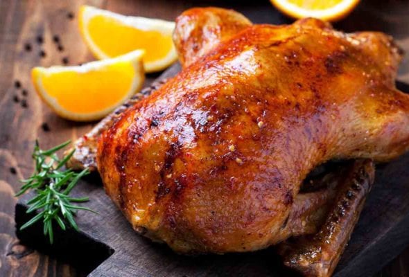 A cooked spit-roasted duck with orange and rosemary in the background.