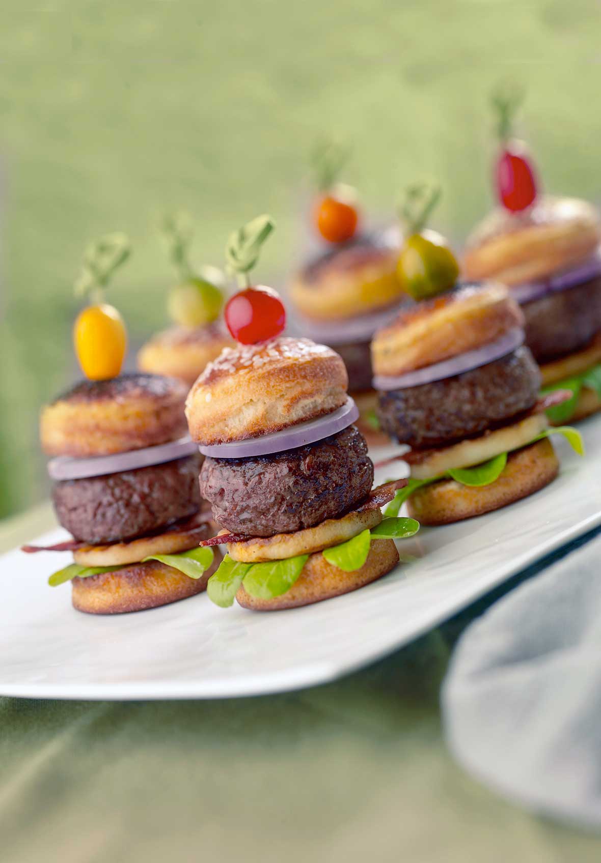 Six blue cheese stuffed bacon sliders, each with a slice of pear, arugula, and a cheery tomato stuck on top