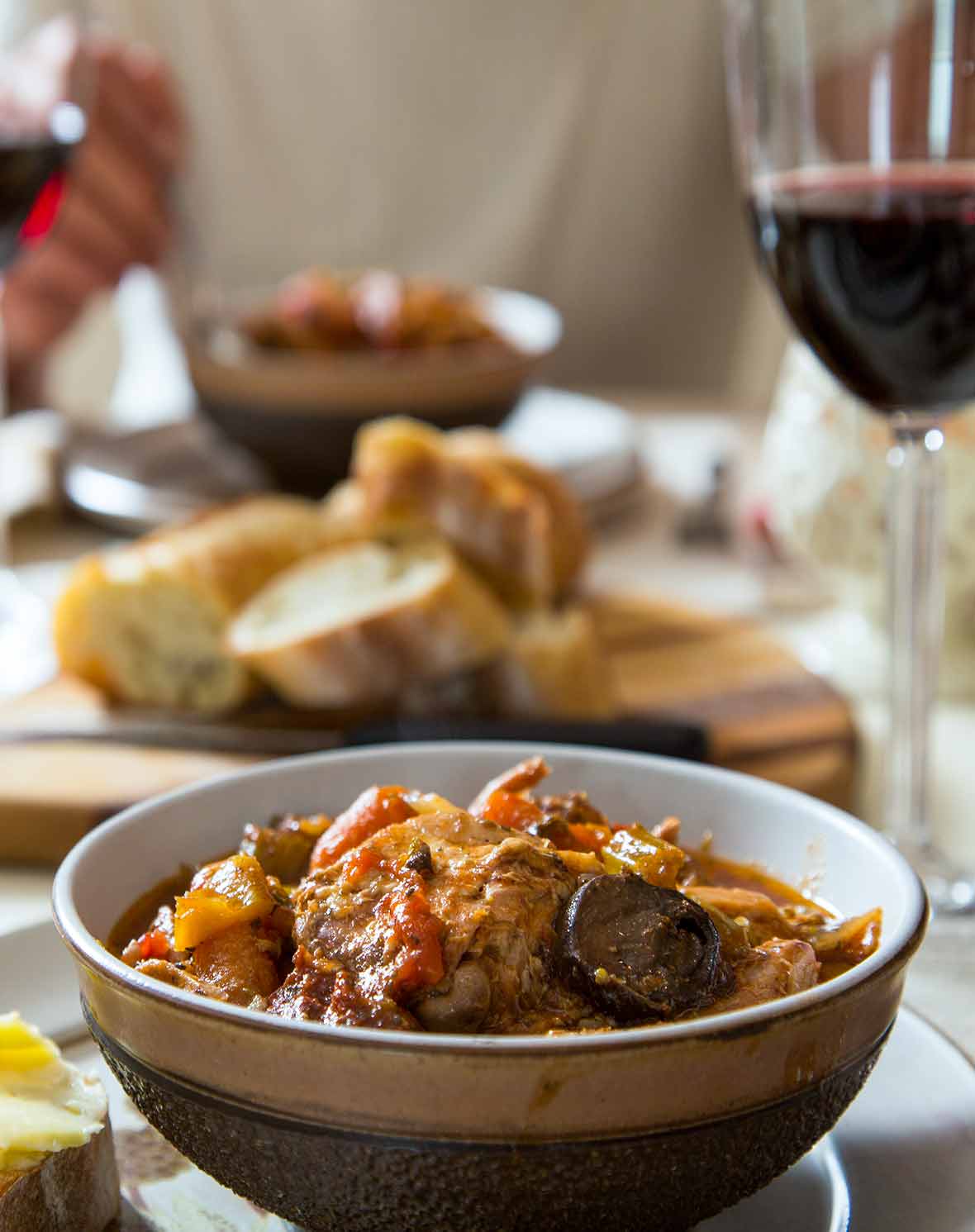 A large bowl filled with Portuguese rabbit hunter style, with a glass of red wine and a cutting board with slice baguette in the background.