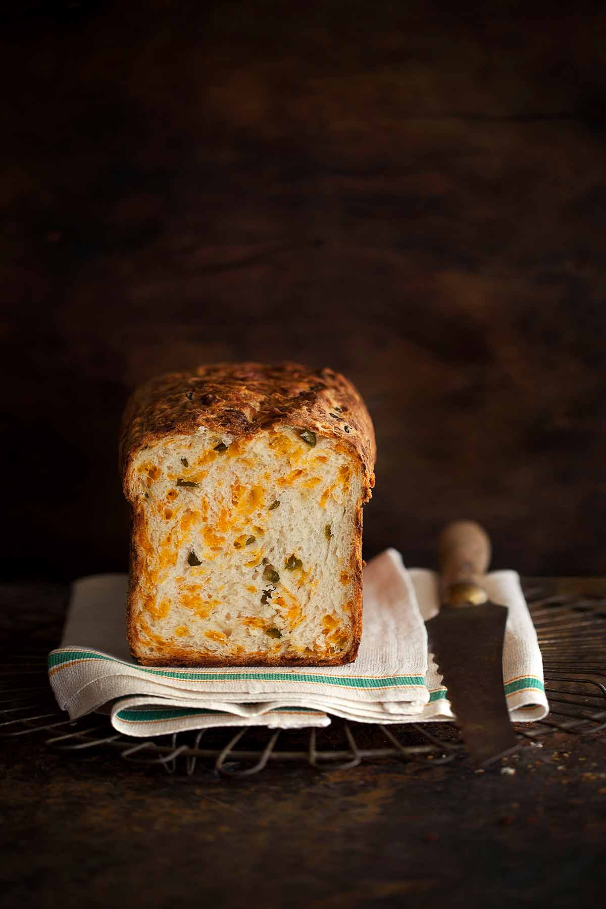 A loaf of no-knead Cheddar-chiles bread on a kitchen towel on a wire rack with two slices cut from it and knife resting beside it.