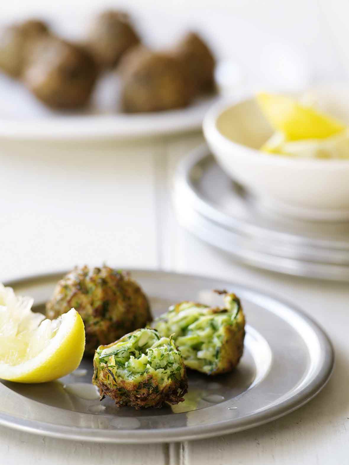 A silver plate with two zucchini fritters, one torn in half, and a wedge of lemon.