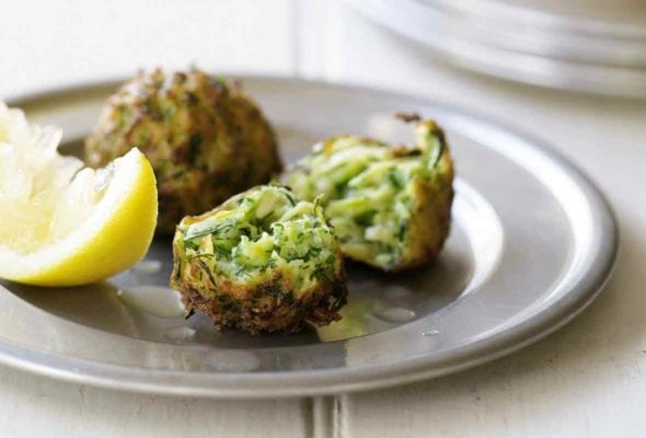 A silver plate with two zucchini fritters, one torn in half, and a wedge of lemon.