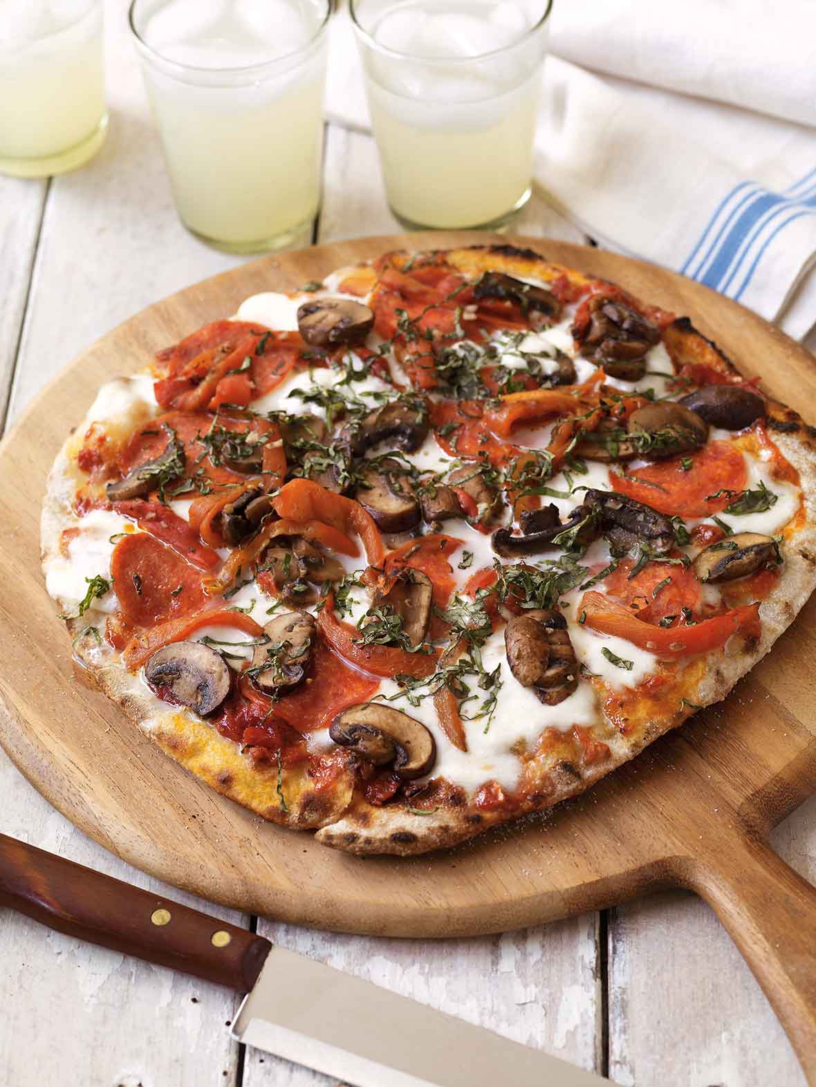 A grilled pizza with pepperoni and mushrooms on a pizza peel with glasses of lemonade, a kitchen towel, and a knife surrounding it.