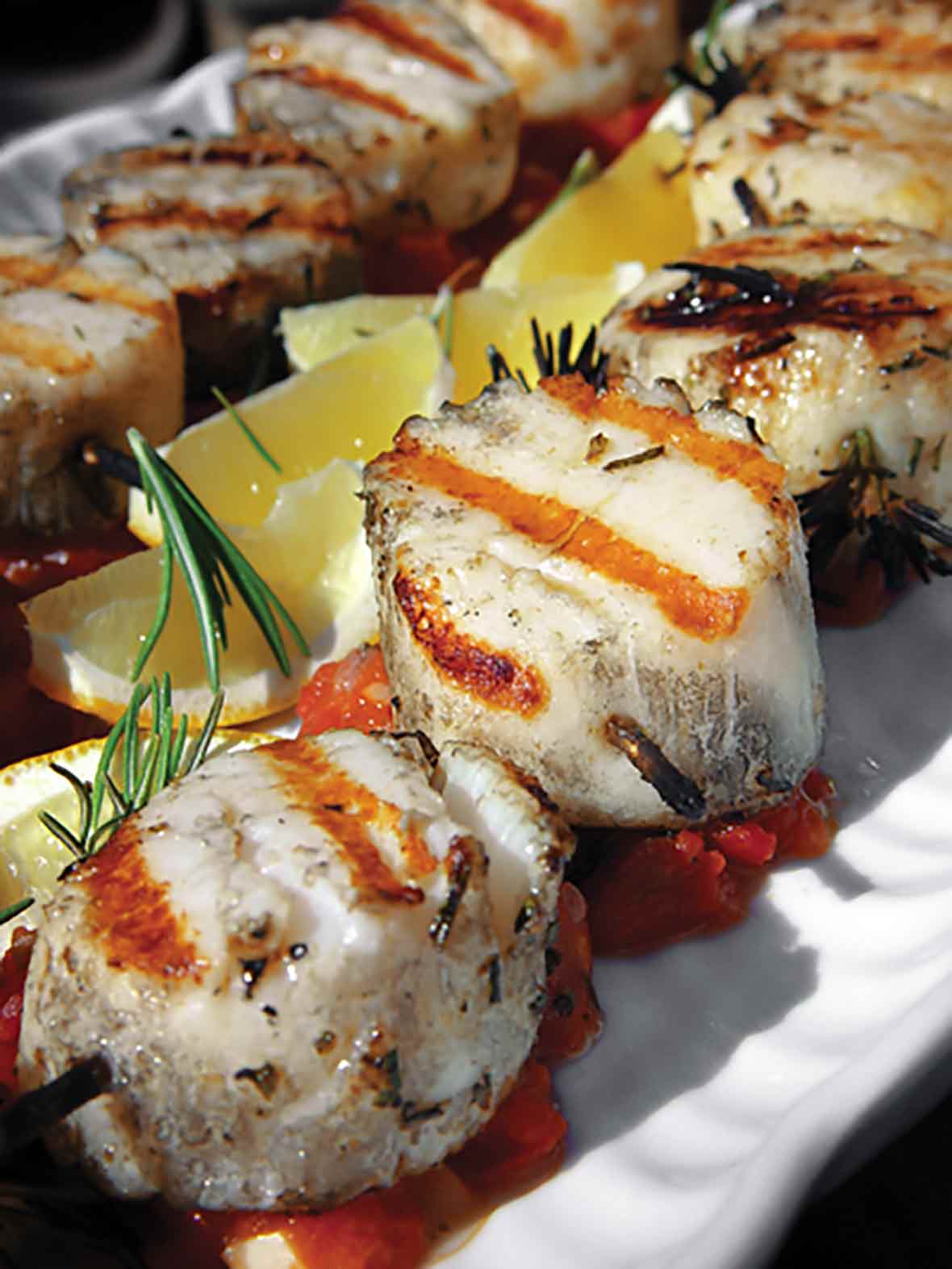 Grilled scallops are threaded on rosemary skewers and sitting on a bed of tomato chutney