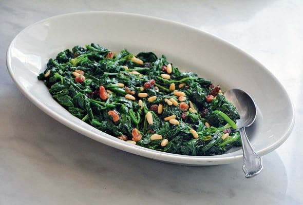 A white oval dish filled with cooked spinach with raisins and pine nuts sprinkled over the top and a spoon resting in the dish.