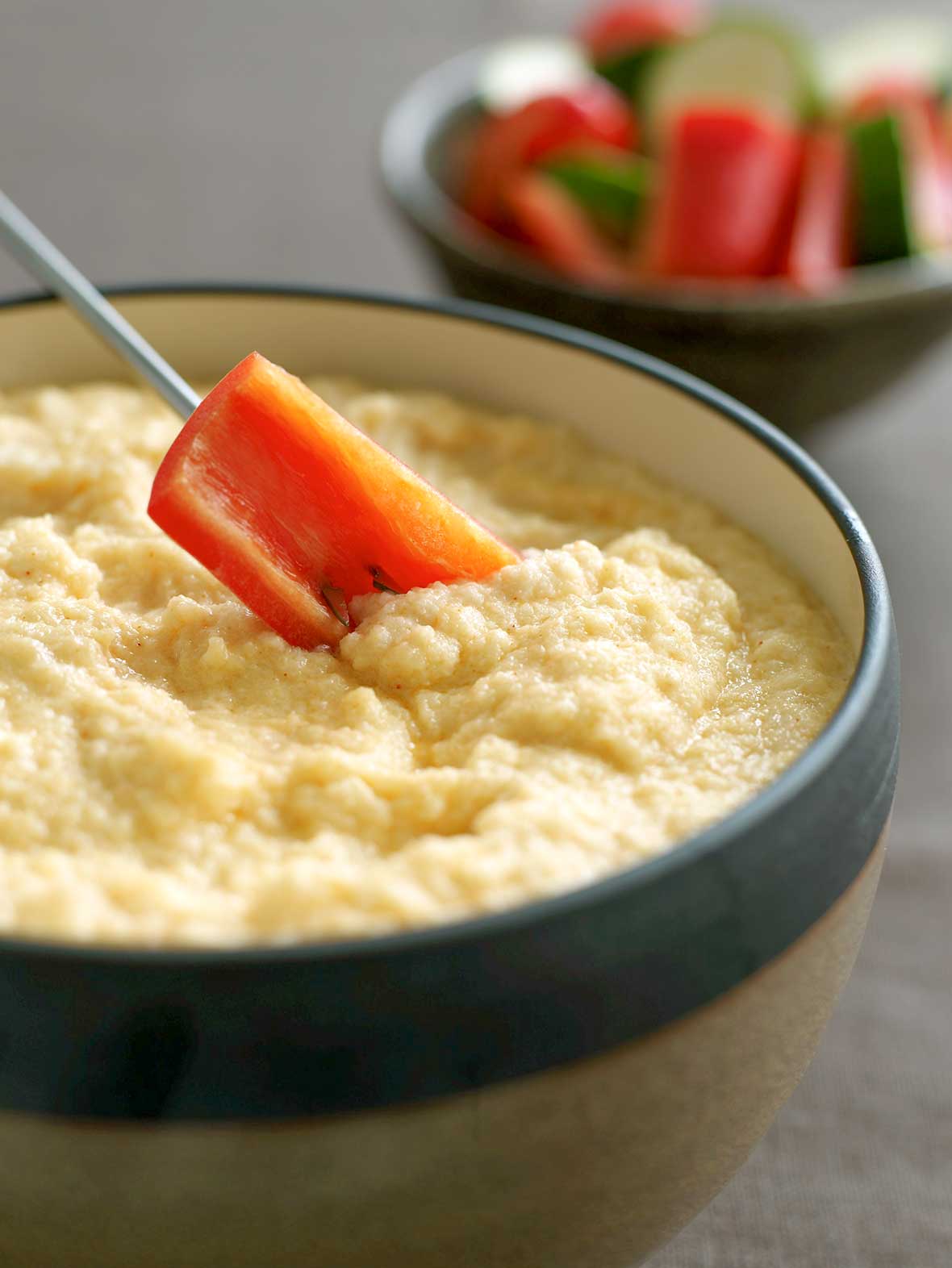 A carrot dipped into A bowl of cauliflower Asiago cheese fondue