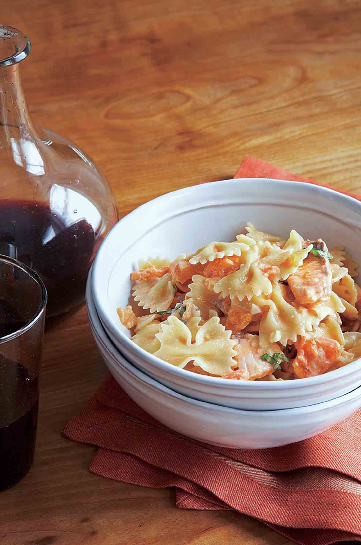 A white bowl filled with farfelle with fresh salmon is nested inside another white bowl with a carafe and glass of red wine in the background.