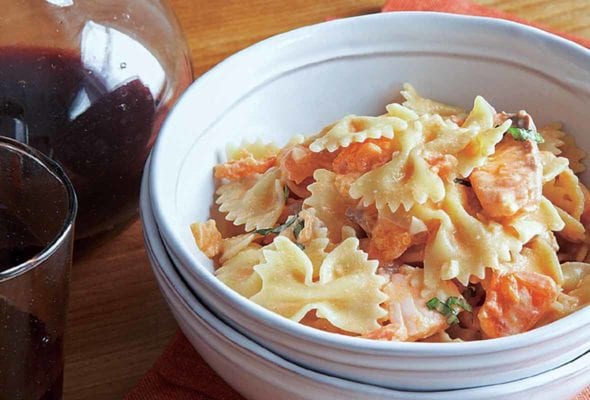 A white bowl filled with farfelle with fresh salmon is nested inside another white bowl with a carafe and glass of red wine in the background.