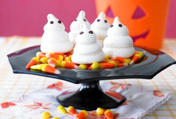 Four meringue ghosts on a bed of candy corn on a black cake stand.