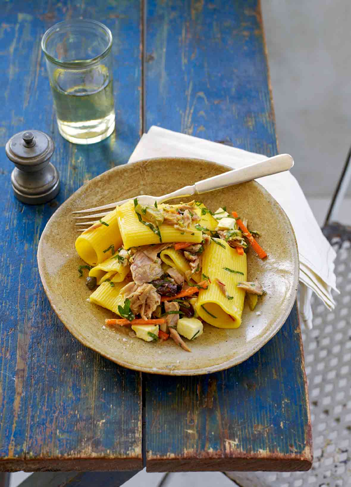 A bowl of saffron pasta salad with tuna, mozzarella, capers, and olives on a blue table