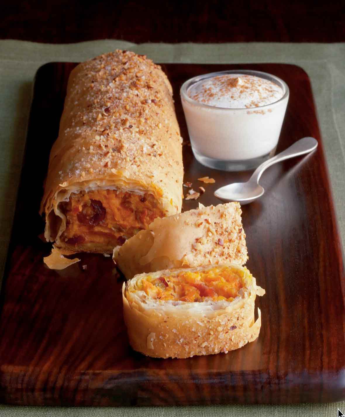 A partially sliced sweet potato and golden raisin strudel, topped with butter and pecans on a wooden board with a bowl of spiced creme fraiche.