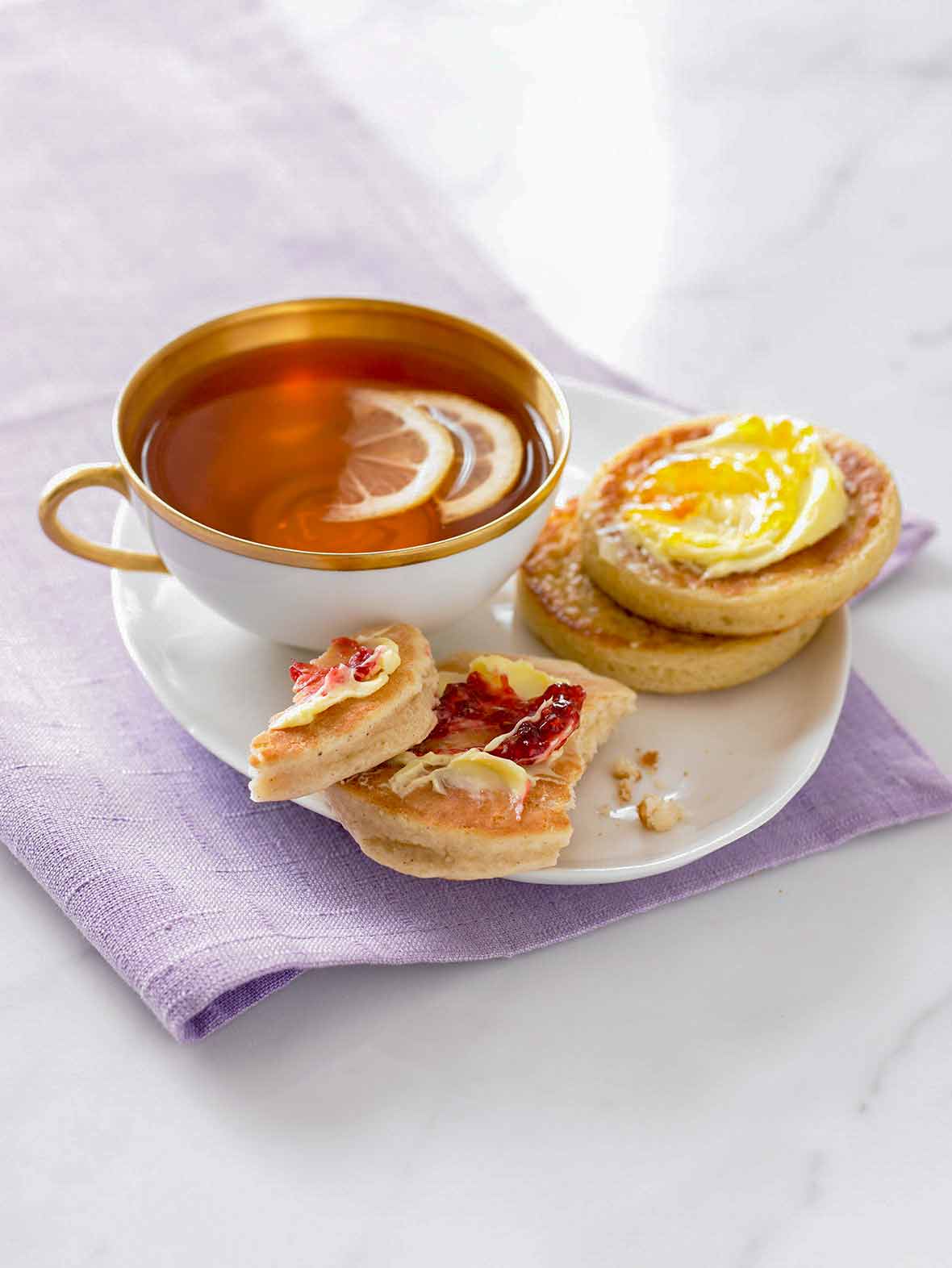 Two English crumpets, one with butter and jam, one with butter and marmalade, on a plate, with a cup of tea nearby