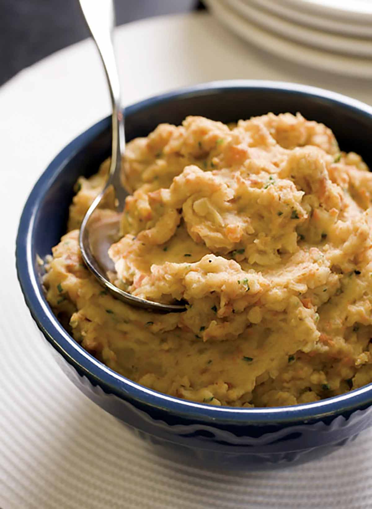 A blue bowl filled with mashed potatoes and root vegetables with a spoon resting inside.
