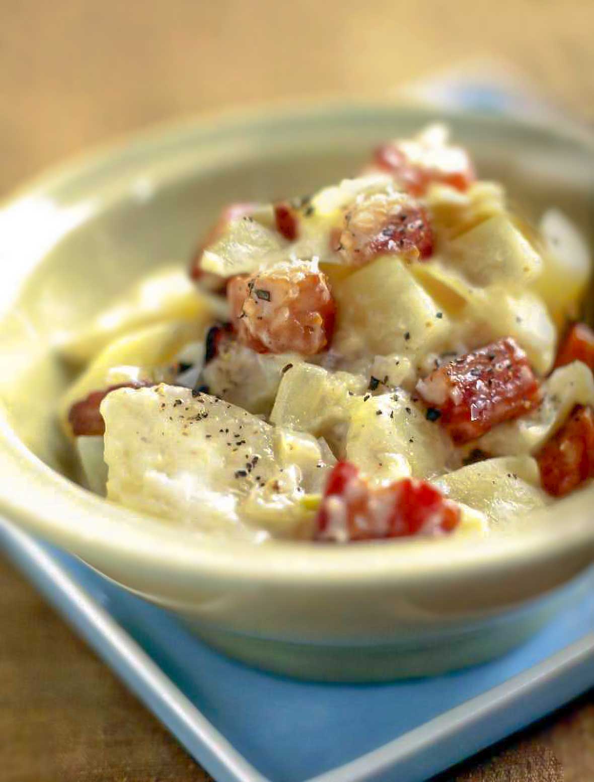 A bowl of apples, pears, potatoes, and bacon that have been braised ; underneath is a blue plate