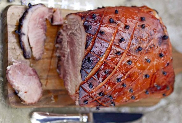 A Barq's root beer-glazed ham on a wooden cutting board with a fork and knife resting beside it.