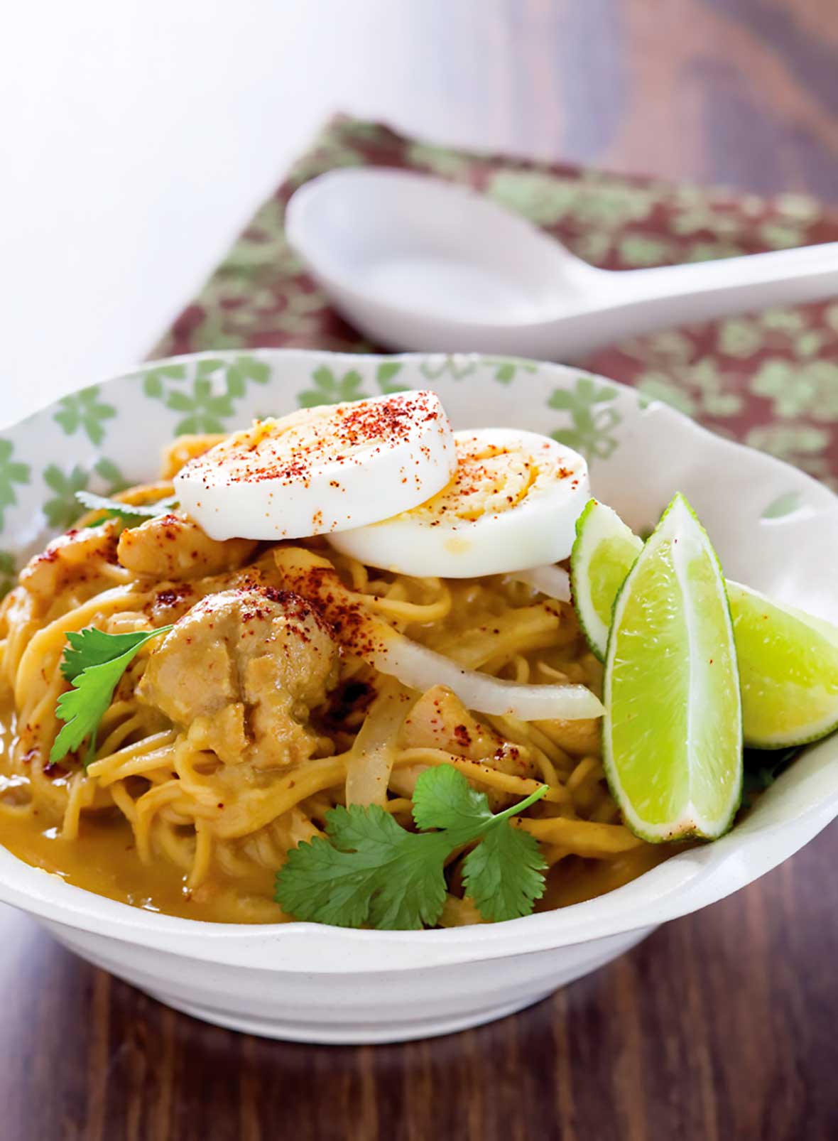 A white bowl filled with chicken coconut noodle soup, topped with cilantro, lime wedges, and hard boiled eggs.