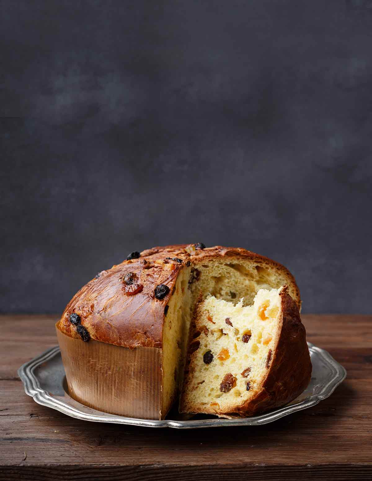 A panettone studded with dried fruit on a silver platter with a couple of wedges cut from it.