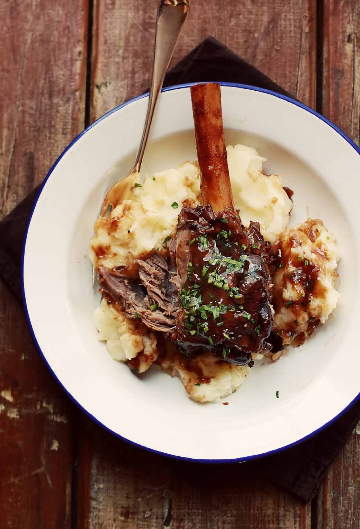 A braised lamb shank in pinot noir, and exposed bone, on mash potatoes, all on a white plate sitting on planks of wood