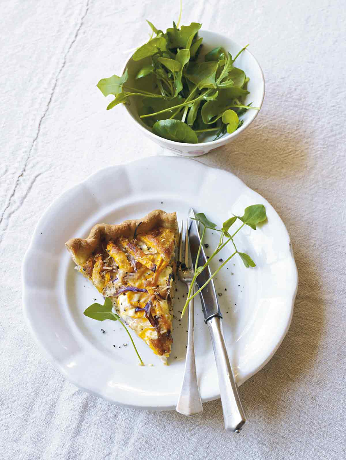 A slice of butternut squash and Parmesan tart on a white plate with a fork and knife, garnished with microgreens.
