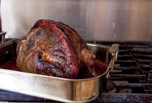 A roasted fresh ham with a maple spice glaze in a metal roasting pan set on a commercial gas range.