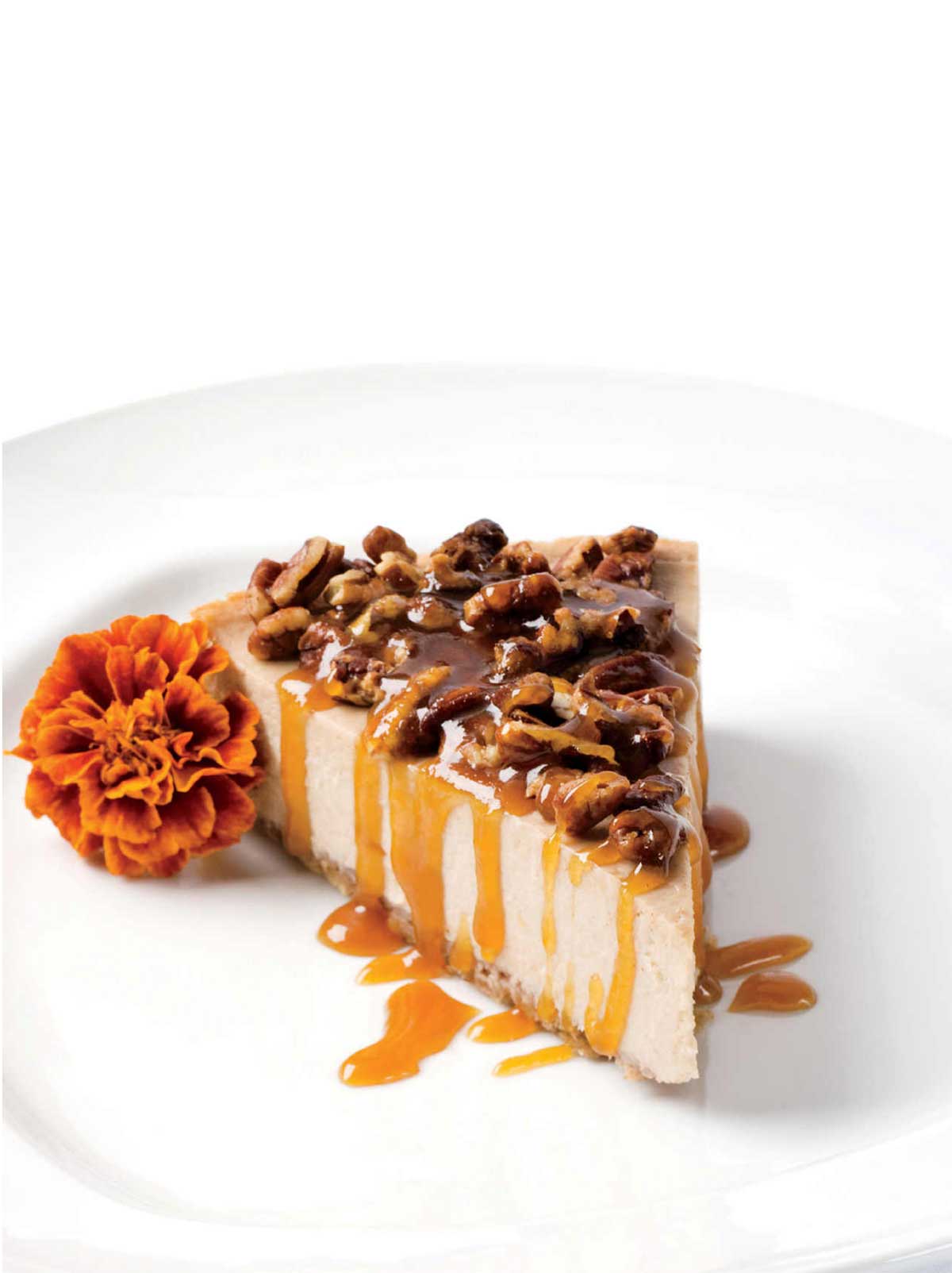 A slice of banana rum cheesecake with maple rum sauce and pecan pieces on top and a marigold beside the cake.